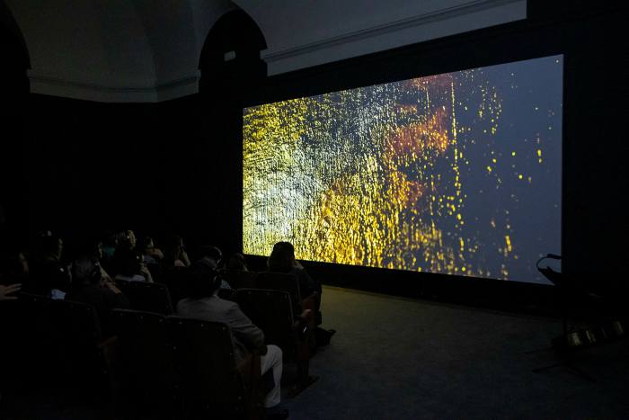 People in a dark room sit and look at a screen displaying a green and yellow abstract image