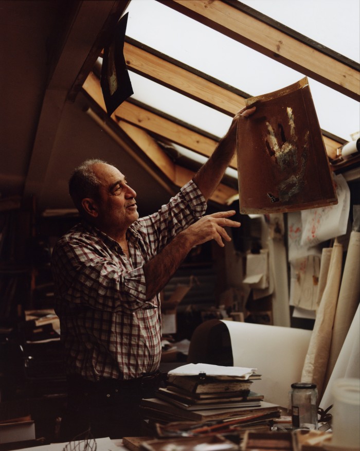 The artist Issam Kourbaj in his studio, holding up a piece of art he has created. It features a large hand