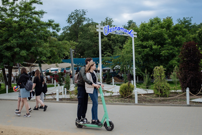 Una pareja joven pasea en scooter por un parque, con una pancarta de Chermorka detrás de ellos