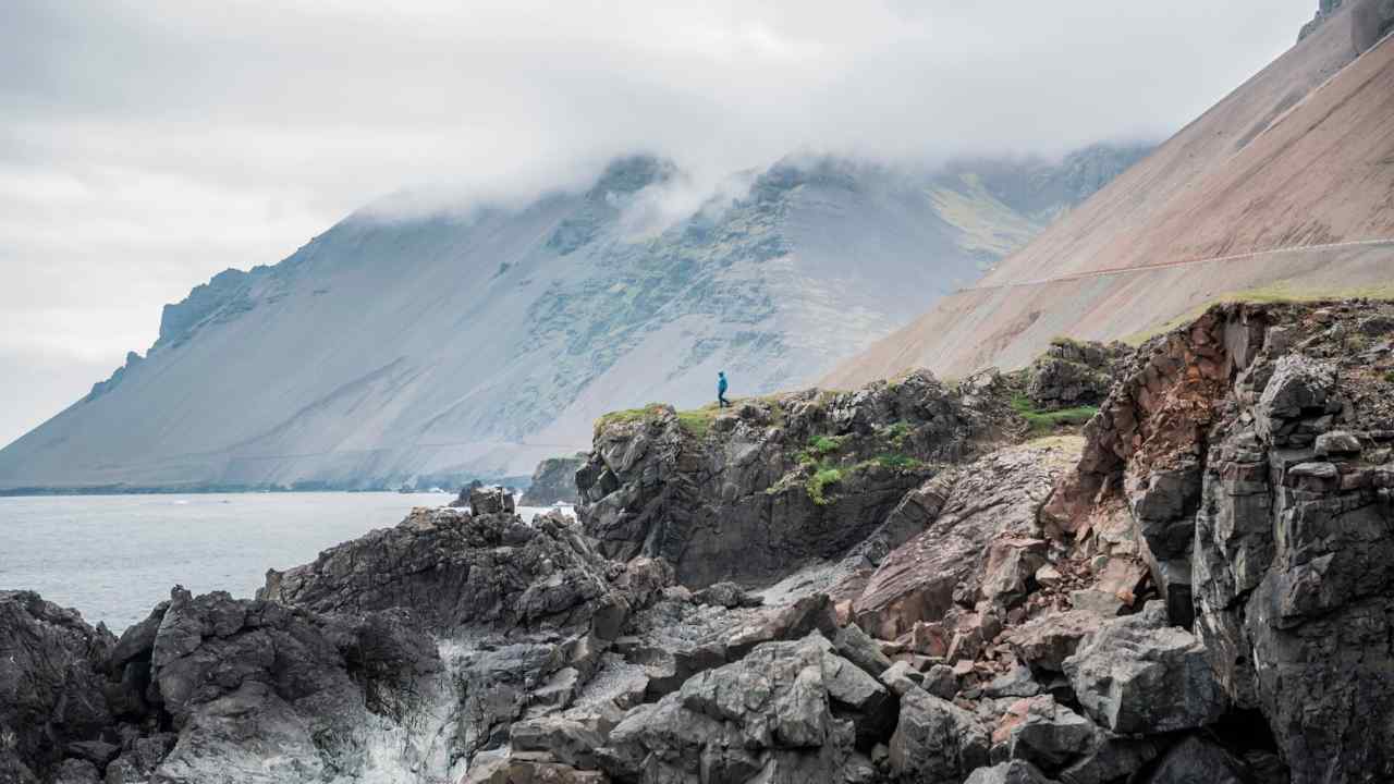 A lone figure strides on rocks by the sea, with mist covering the tops of mountains in the distance