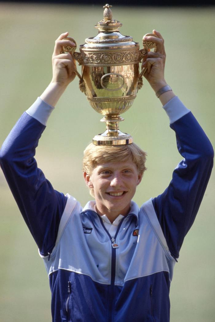A smiling Boris Becker stands smiling, holding a gold trophy on his head