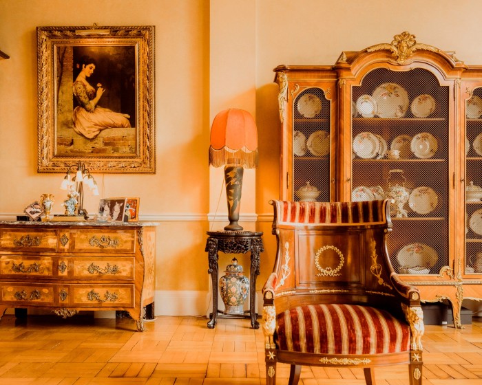 A room with a large ornate cabinet full of china, a striped armchair and a gilt-framed painting of a woman in Spanish dress