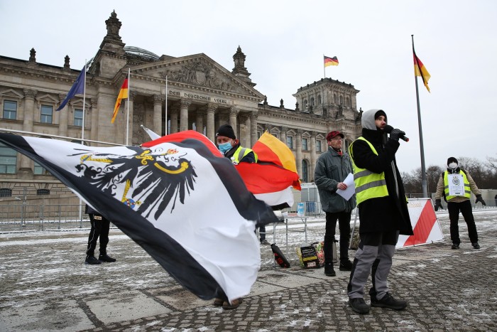 Three people in winter coats and holding a large flag stand in front of the Reichstag building in Berlin
