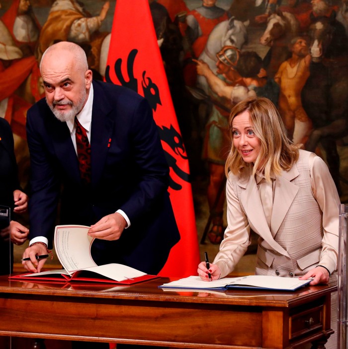 A man and woman sign documents on a table. There is an Albanian flag in the background
