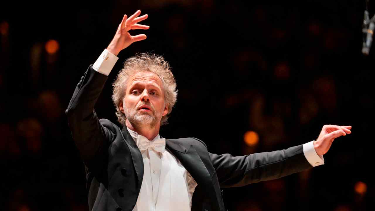 A man with wild-looking greying hair, wearing white tie, conducts an orchestra