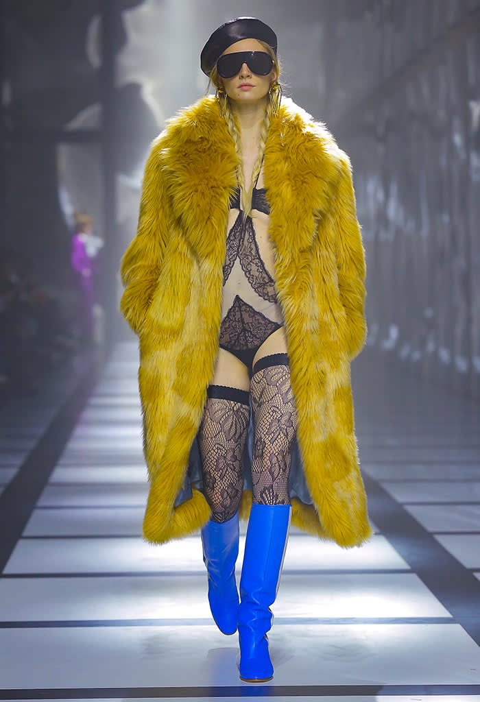 A female model in beret and sunglasses wears black lace underwear under a bold yellow fur coat with blue high boots