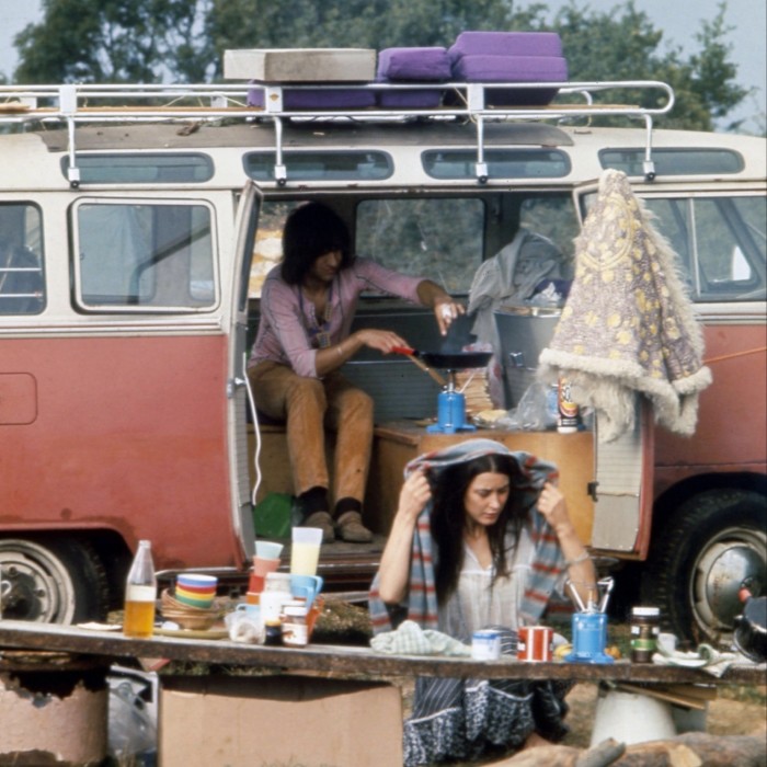 A man sits inside a campervan, cooking over the stove, while a woman sits outside in the rain, holding a scarf over her head 