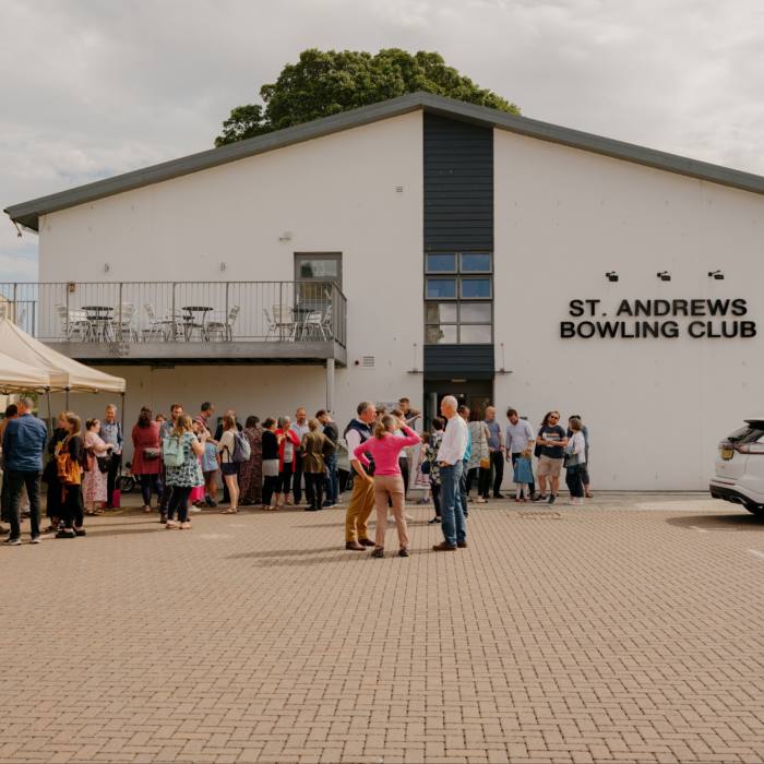People stand outside a hall which has a sign reading: St Andrews Bowling Clu
