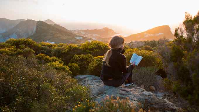 A woman sits on a rocky hillside, reading in daylight