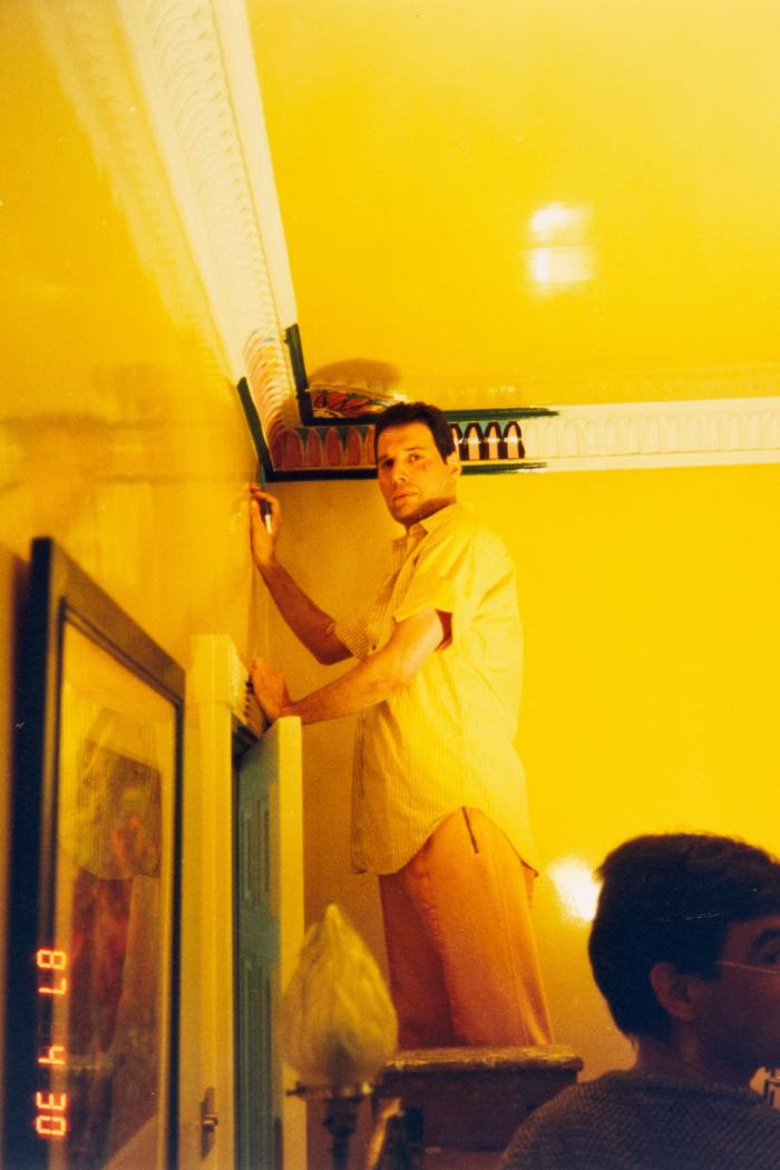 Freddie Mercury stands on a stepladder, painting the cornice of a bright yellow room