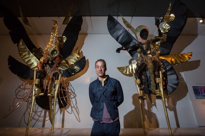 A man stands in front of two large metallic sculptures which resemble winged creatures