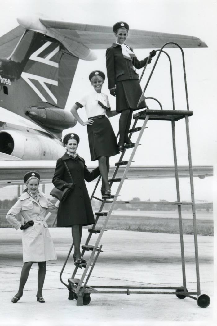 Three air stewardesses stands on aircraft steps near the tail of a plane. A fourth stands on the ground