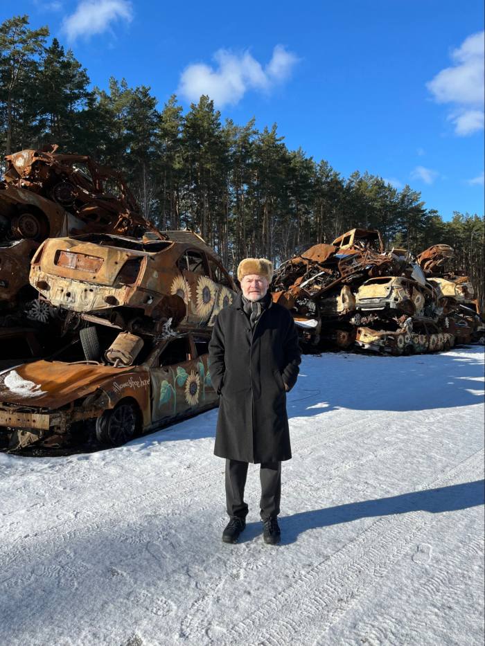 A man stands in front of a scrapheap of rusting and ruined cars