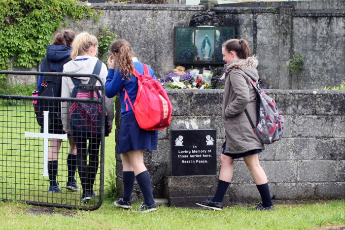 The four schoolgirls pass through a gate with a white cross, towards a statuette of the Virgin Mary surrounded by flowers. A plaque reads: ‘In loving memory of those buried here. Rest in peace’