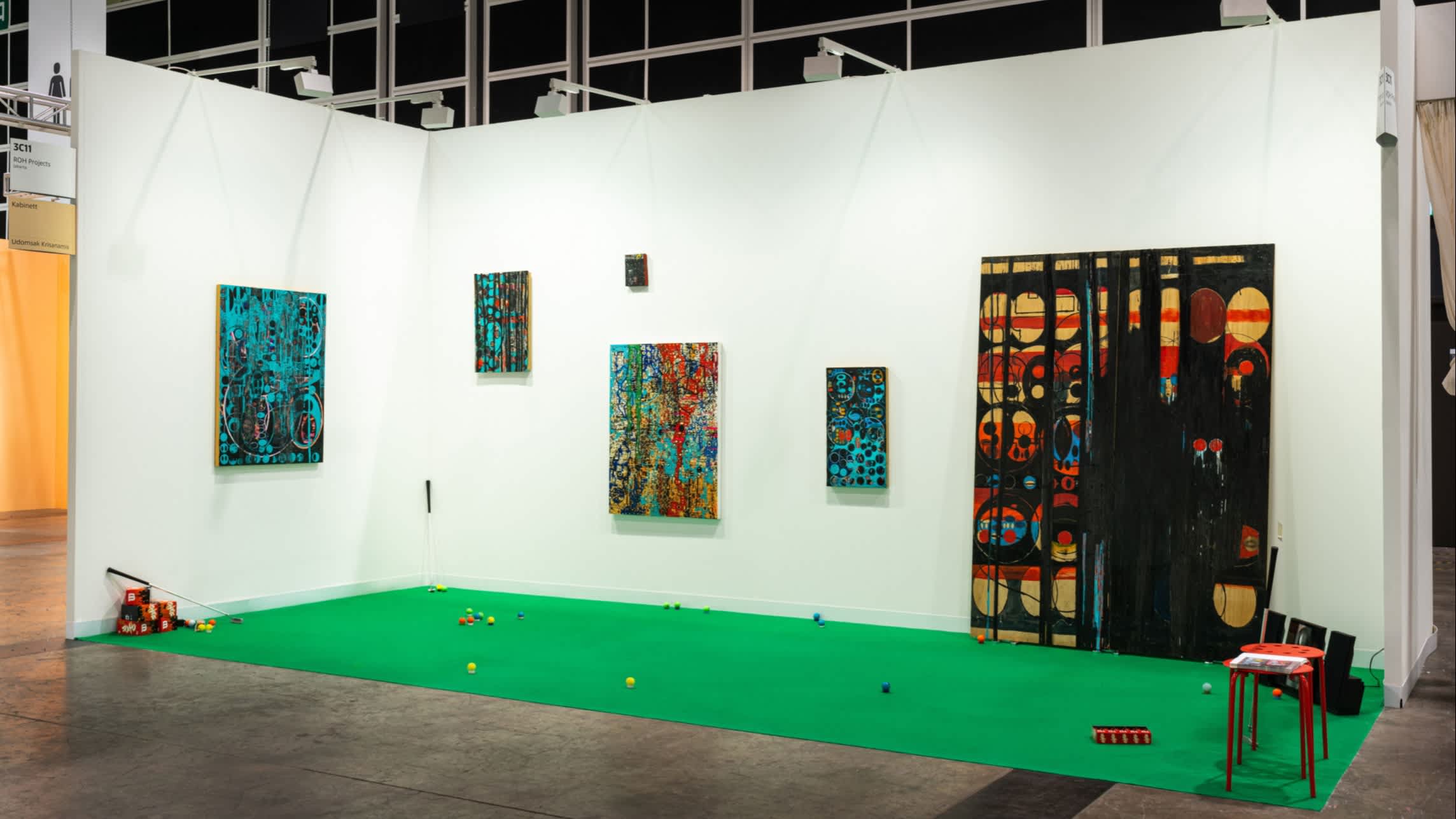 An art installation consisting of a fake corner of an art gallery, with white walls and paintings, and a green carpet with coloured balls and a golf club