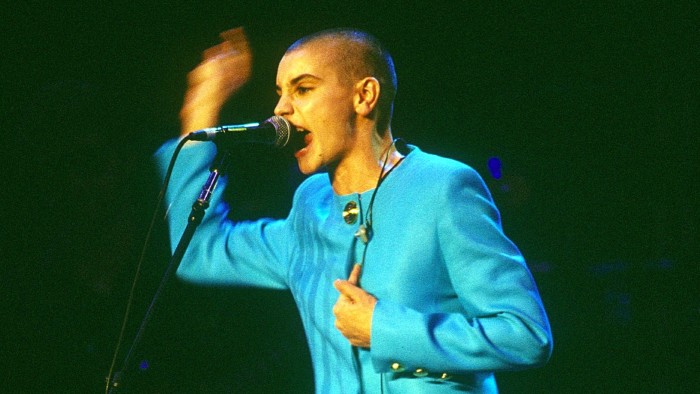 Sinéad O’Connor stands angrily at a microphone, gesturing with one hand 