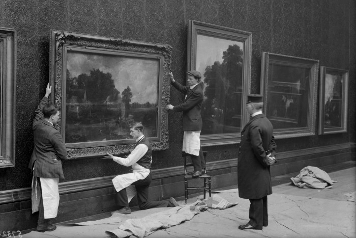 Three young men hold up a large framed painting against the gallery wall as a guard looks on 