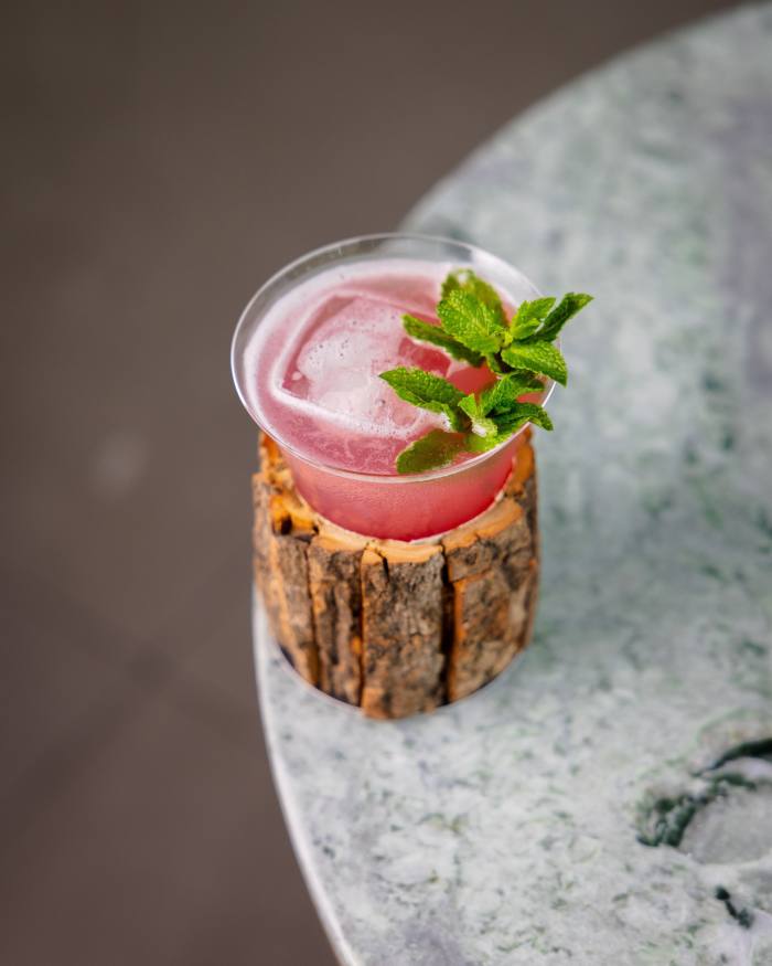 Popinjays' Cotton Tree Cocktail - een donkerroze drankje in een klein driehoekig glas dat in een ruwe houten container wordt bewaard