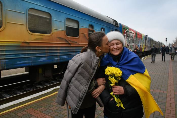 On a station platform, a young woman kisses and older woman who holds a bunch of yellow flowers