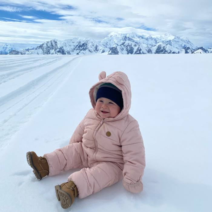 A toddler in a padded all-in-one suit sits smiling on the ice