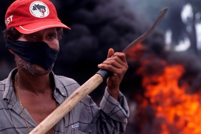Un hombre con una máscara negra y un sombrero del MST sostiene una hoz con humo y fuego detrás de él