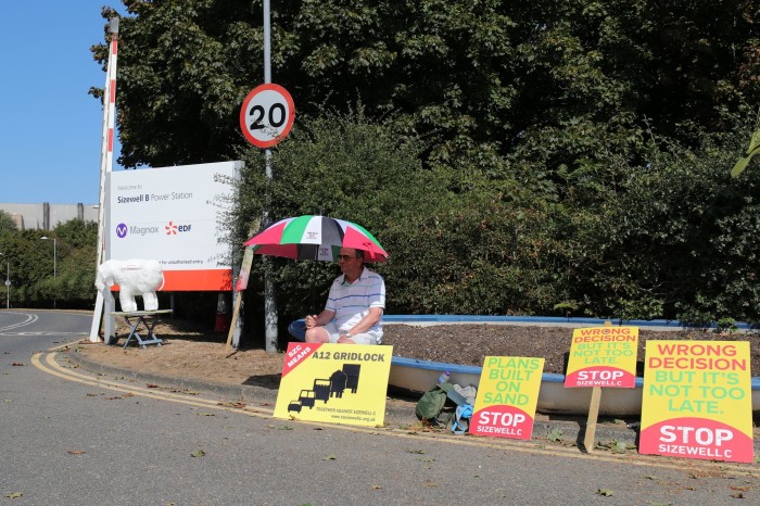 A lone demonstrator with multiple placards and an umbrella to protect from the sun sits  outside Sizewell B