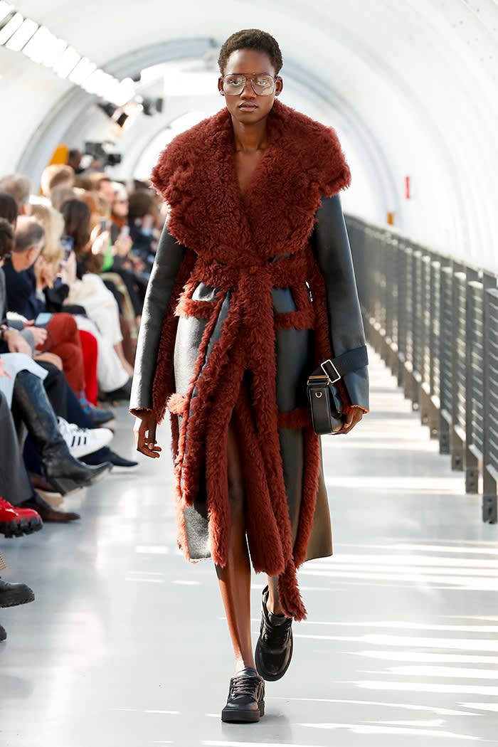 A female model on the catwalk in a long coat adorned with bulky red fur collar and trim