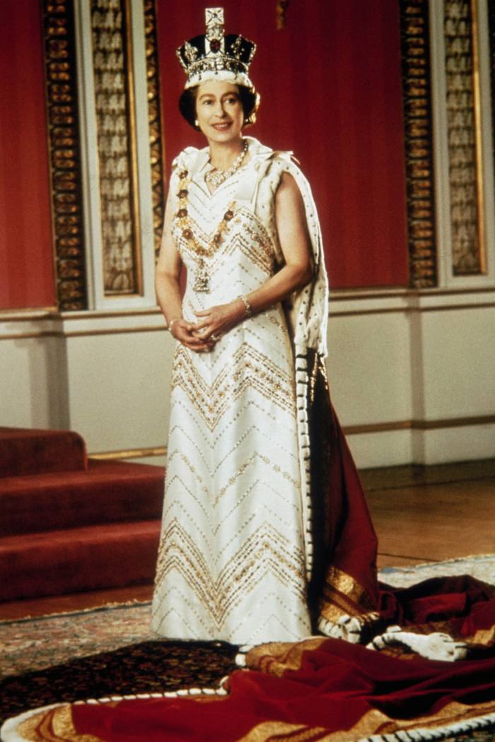 La reine photographiée en 1977. Elle est debout souriante et vêtue d'une longue robe, d'un manteau fluide et d'une couronne