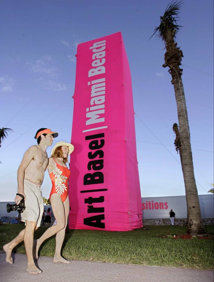 A man and a woman in swimsuits walk past a pink Art Basel advertising column jutting out from the edge of the grass 