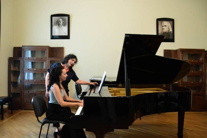 A young woman sits playing a piano, while an older woman leans over her and points to the sheet music