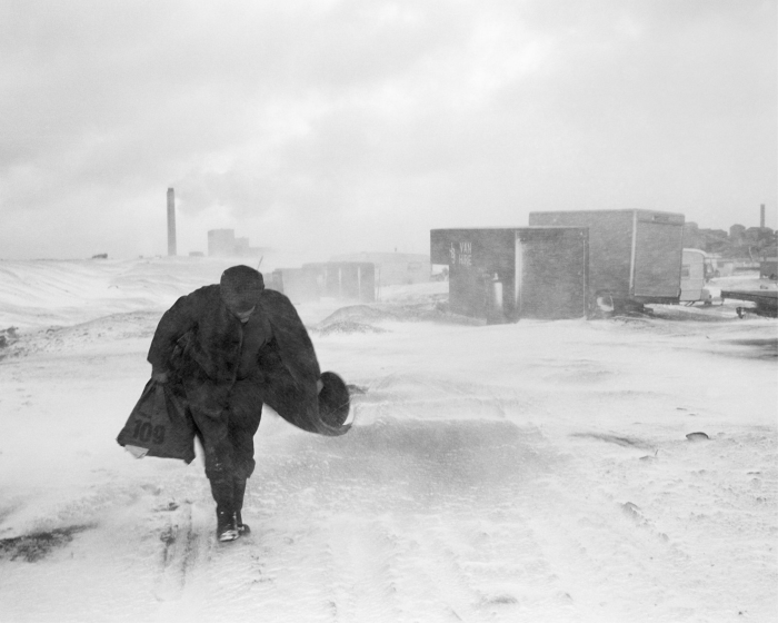 رجل يمشي في الثلج ، متجهًا نحو الكاميرا ، خلفه شاحنات وحاويات ومداخن