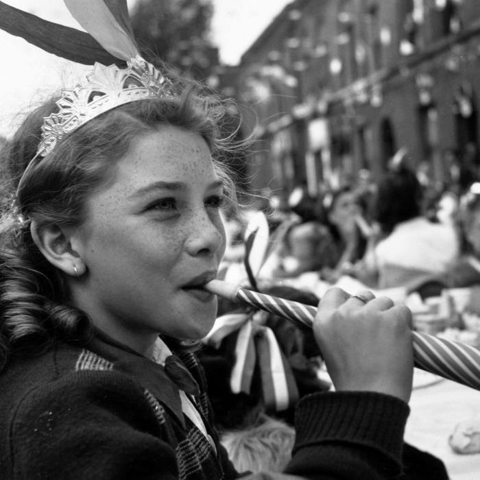 A young girl wearing a paper hat blows a paper trumpet at an outdoor street party