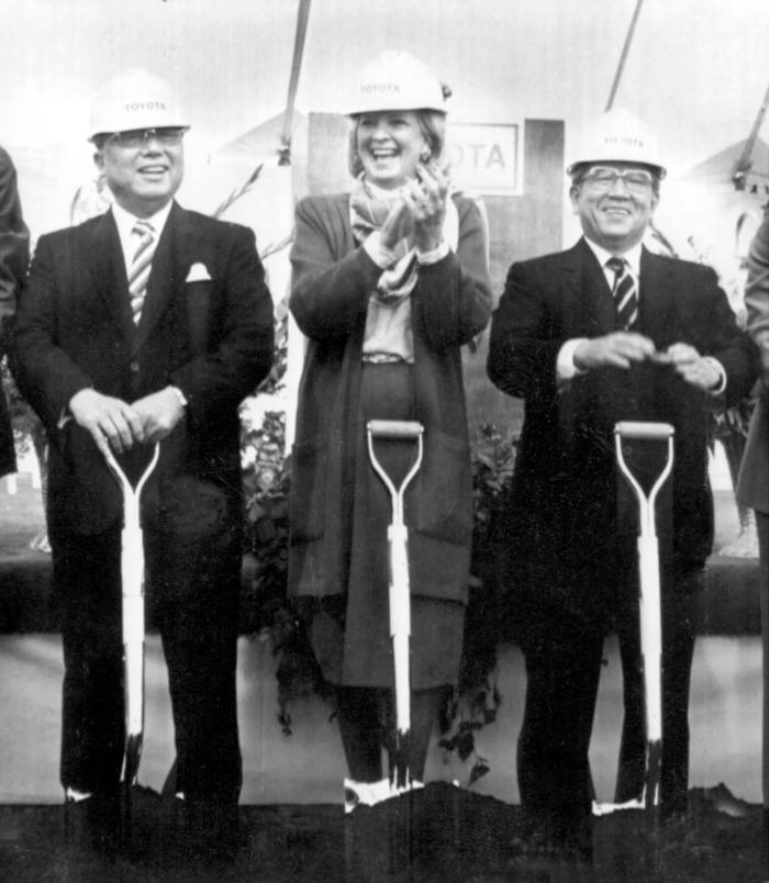 A helmeted woman is flanked by two men in suits holding shovels on fresh dirt.