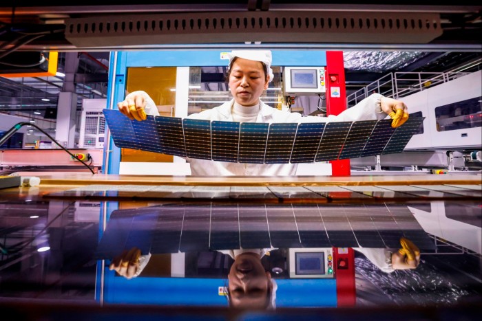 A worker in a cleanroom suit inspects a large flexible solar panel in a high-tech manufacturing setting, with the panel’s reflection visible on a shiny surface below