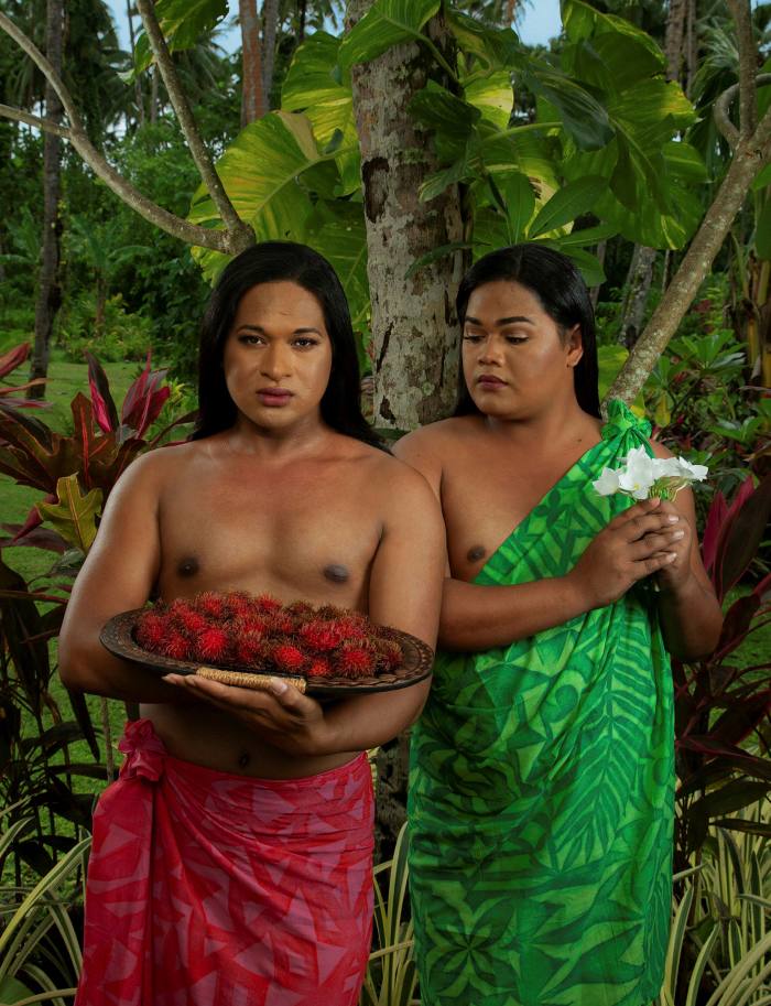 Two people stood standing side by side, behind them is a jungle. One is looking at the viewer and has a red sheet tied around their waist and holds a platter of exotic red fruits; the other wears a green sheet draping down from their left shoulder and holds white flowers in their hands. Their gaze is looking down at the platter