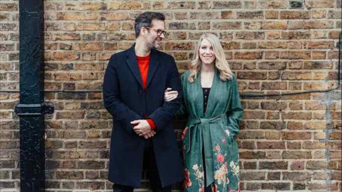 A tall man in a dark overcoat looks smilingly at a blond woman in a green coat who has her arm through his