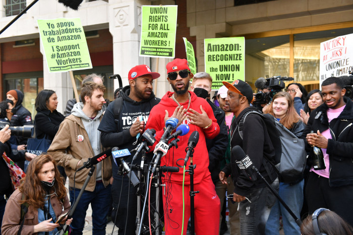 Amazon workers with placards stand around union organiser Christian Smalls, who is wearing a red tracksuit, peaked cap and darkglasses, as he speak to the press