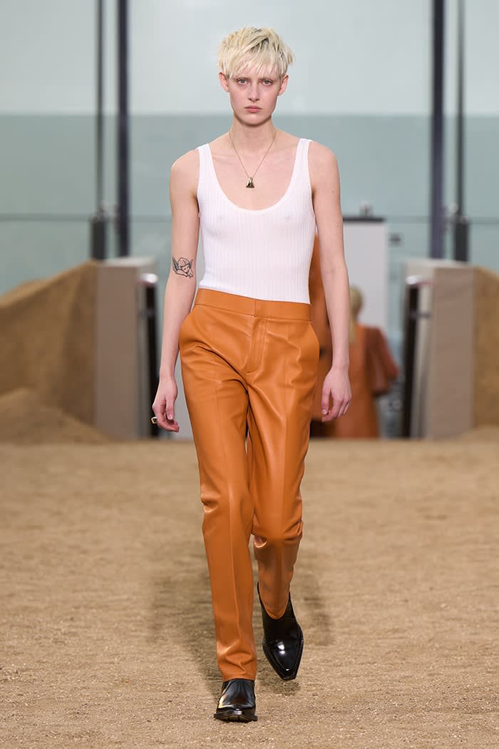 An androgynous female model with spike blond hair wearing a sleeveless white top and orange trousers