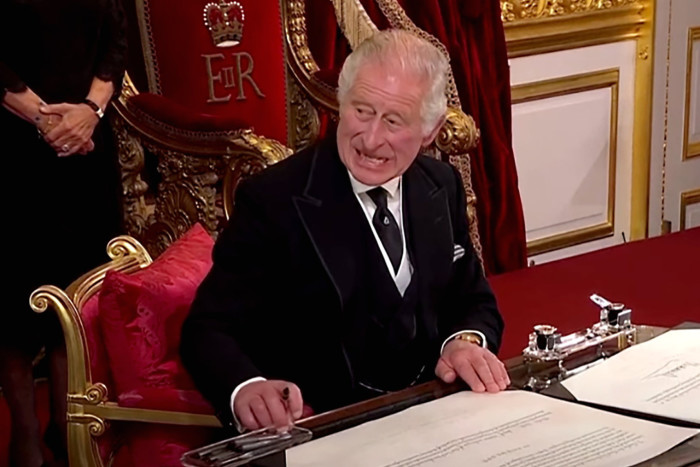 El rey Carlos III, sentado en un escritorio, hace un gesto a los asistentes para que retiren el bolígrafo en la esquina del escritorio.