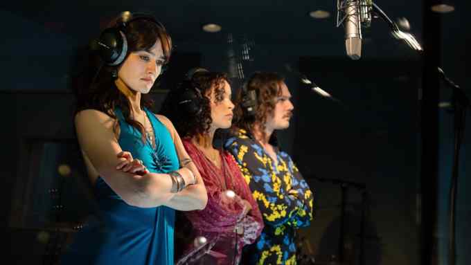 Two women and a man wearing 1970s clothing stand in a line in a recording studio with a microphone suspended in front of them; all look disgruntled and have their arms folded