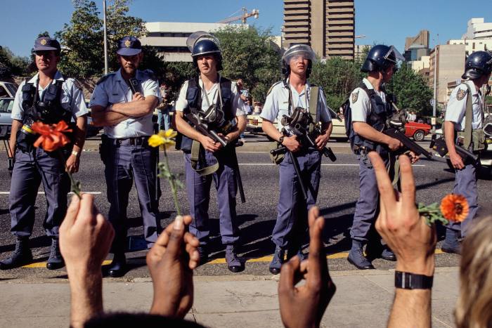 Riot police block anti-apartheid protesters who are holding flowers and flashing peace signs