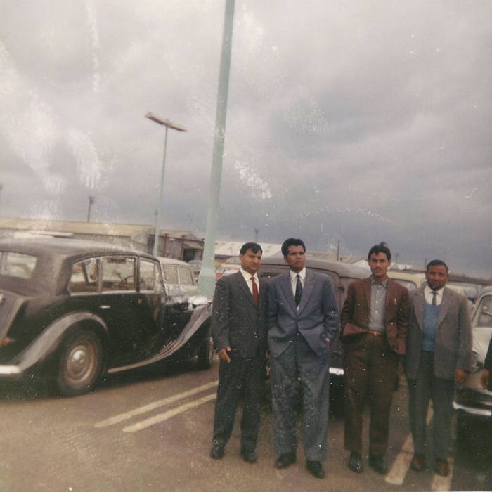 Four young men in suits pose for a photo in 1963, standing next to a large parked car