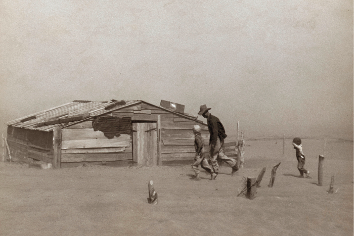 A man and two small boys walk through a thick cloud of dust towards a ramshackle house