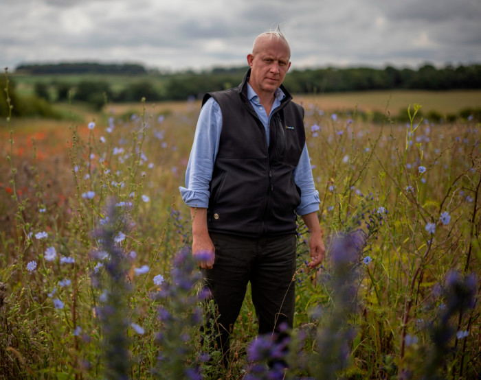 Jake Fiennes, wearing jeans, light blue long-sleeved shirt and black bodywarmer, stands in a meadow of wild flowers with row trees in the distance 