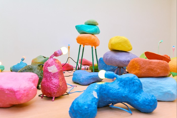 Una vista de instalación de coloridos objetos escultóricos, algunos con accesorios de iluminación adjuntos, dispuestos aparentemente al azar en el piso de un espacio de galería.