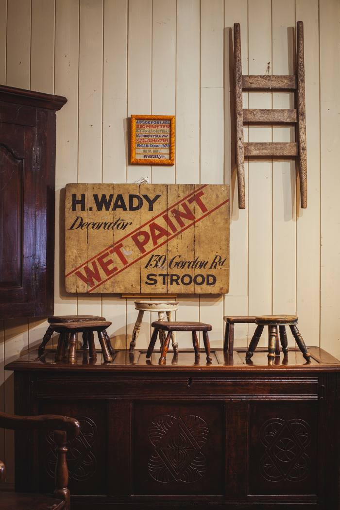 An early-20th-century “Wet Paint” sign, £575. Above it is a small Welsh sampler dated 1873, £350, and a rare Welsh hand-barrow, £345. Small 19th-century Welsh stools and candle stands, from £135, stand on a carved 17th-century Welsh oak chest, £1,680