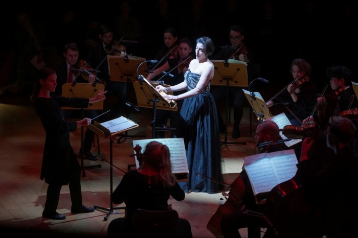 A female singer in a black ballgown stands amid an orchestra