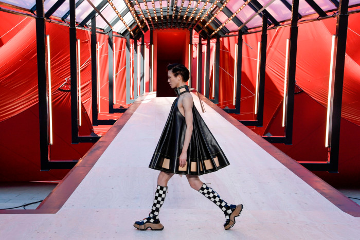 A catwalk model walks across a stage wearing a sleeveless dress of pleated black material and long checkerboard pattern socks