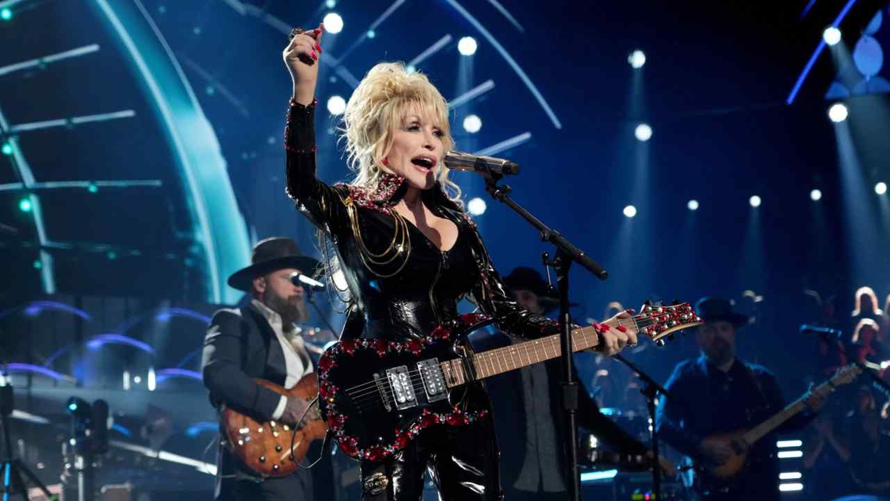 Dolly Parton stands holding an electric guitar behind a microphone stand