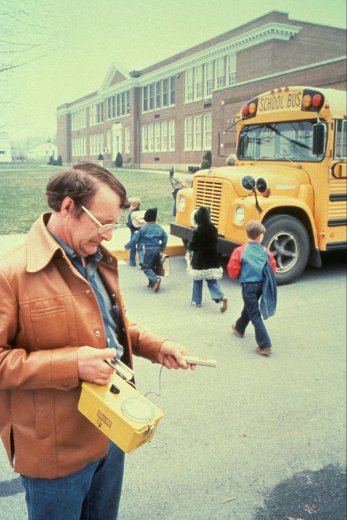 A man in a 1970s-style leather jacket checks the reading on an instrument in a yellow casing. Behind him, children cross the road towards a yellow school bus